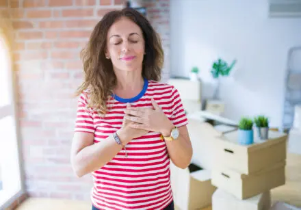 A woman in her 40s with her eyes closed, breathing, with her hands over her heart. She has wavy, shoulder-length brown hair, and is wearing a red and white horizontally striped t-shirt. She is standing in a well-lit room. An office with a desk and boxes is blurred out in the background, as well as an arched door with a red brick wall.