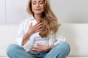 Photo of middle-aged woman meditating. She had one hand on her chest and another on her tummy. She has long light brown hair, is wearing an open white blouse over a white top, and light blue jeans. It's a bright room with a white background behind her.