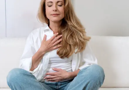 Photo of middle-aged woman meditating. She had one hand on her chest and another on her tummy. She has long light brown hair, is wearing an open white blouse over a white top, and light blue jeans. It's a bright room with a white background behind her.