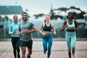 4 people in their 20s and 30s running, coming towards the camera. There's a man slightly in front and three women just behind - one to the left of the man and two to the right of him, from camera perspective. They're all wearing sports gear and looking happy.