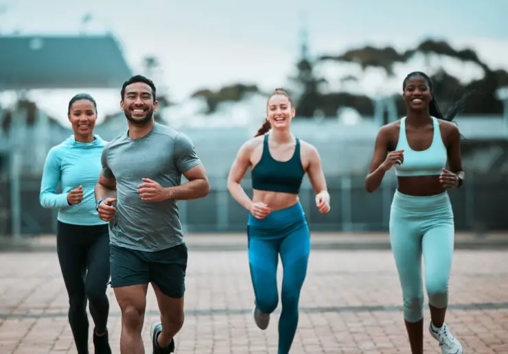 4 people in their 20s and 30s running, coming towards the camera. There's a man slightly in front and three women just behind - one to the left of the man and two to the right of him, from camera perspective. They're all wearing sports gear and looking happy.