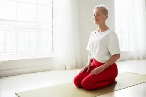 Middle aged woman in a kneeling-meditation pose in a brightly lit studio. She is wearing red leggings and a white t-shirt. There are two windows behind her with bright sunshine shining through them, with white wispy curtains open, illuminating the room. She is kneeling on an olive green mat, which sits on a wooden floor.