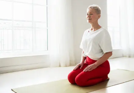 Middle aged woman in a kneeling-meditation pose in a brightly lit studio. She is wearing red leggings and a white t-shirt. There are two windows behind her with bright sunshine shining through them, with white wispy curtains open, illuminating the room. She is kneeling on an olive green mat, which sits on a wooden floor.