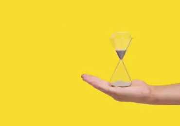 Photograph of a hand with open palm, with an hourglass sitting on it. It's run less than half way through. The photo is set against a bright yellow background.