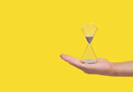 Photograph of a hand with open palm, with an hourglass sitting on it. It's run less than half way through. The photo is set against a bright yellow background.