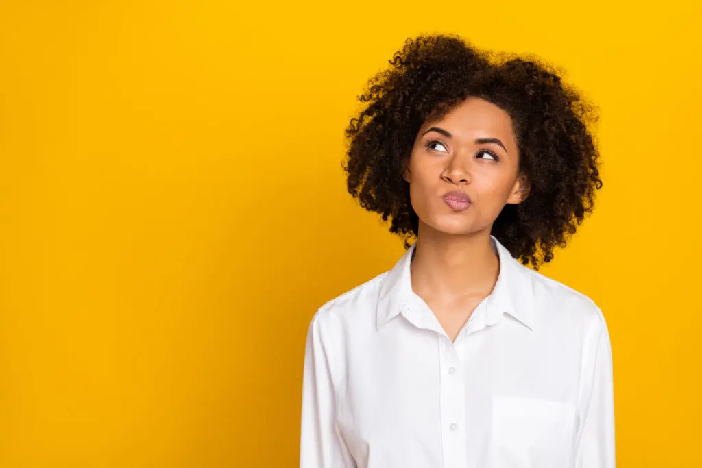 Girl in her 30s with dark wavy hair making a curious face and looking up with her eyes and closed mouth to the top right. She has her lips pursed and pointing them up in the same direction as her eyes. She is wearing a while blouse and the background is a bright mustard-yellow.