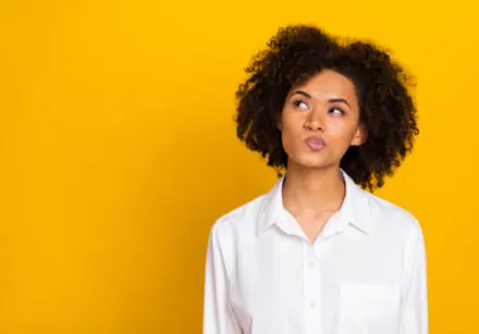Girl in her 30s with dark wavy hair making a curious face and looking up with her eyes and closed mouth to the top right. She has her lips pursed and pointing them up in the same direction as her eyes. She is wearing a while blouse and the background is a bright mustard-yellow.