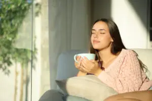 Young woman relaxing at home drinking tea from a large pale blue cup. She sits at the window and the sun is shining. Her eyes are closed as she savours the quiet. She has long, dark brown hair, and is wearing a peach coloured top. She is sitting on a comfy sofa.