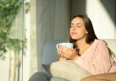 Young woman relaxing at home drinking tea from a large pale blue cup. She sits at the window and the sun is shining. Her eyes are closed as she savours the quiet. She has long, dark brown hair, and is wearing a peach coloured top. She is sitting on a comfy sofa.