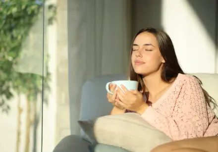 Young woman relaxing at home drinking tea from a large pale blue cup. She sits at the window and the sun is shining. Her eyes are closed as she savours the quiet. She has long, dark brown hair, and is wearing a peach coloured top. She is sitting on a comfy sofa.