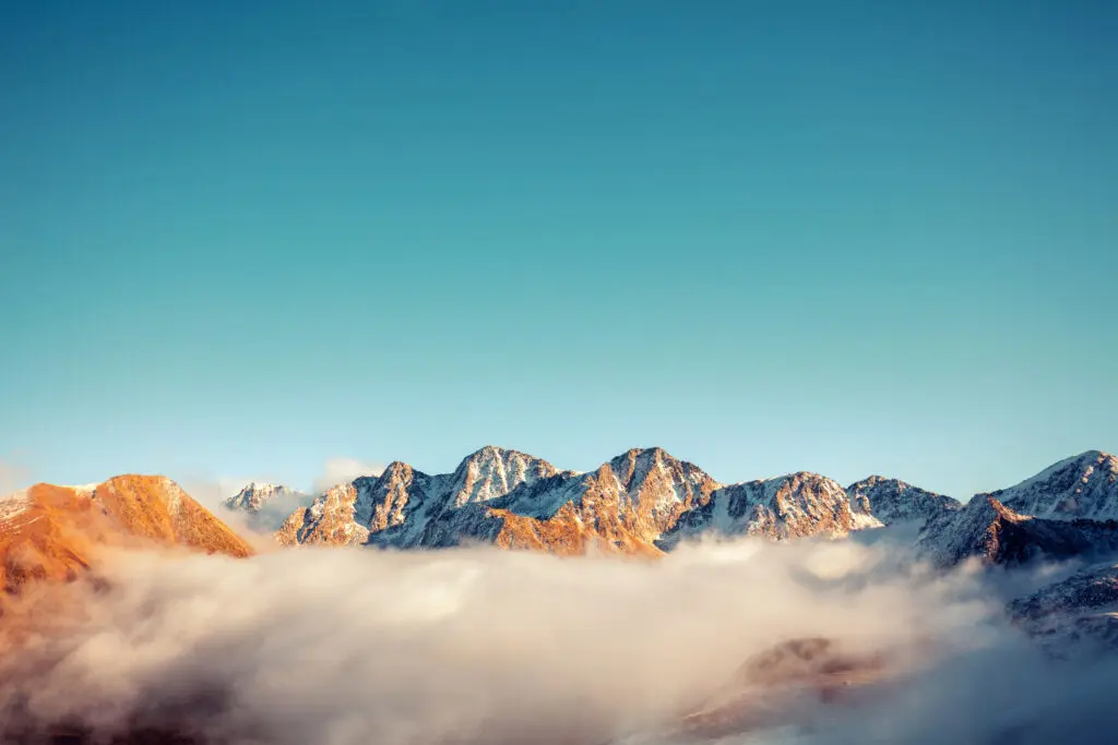 partially snow-capped mountain ranger, looking over the top of a canopy of clouds. The sky is light blue in the background.