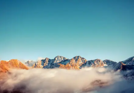 partially snow-capped mountain ranger, looking over the top of a canopy of clouds. The sky is light blue in the background.