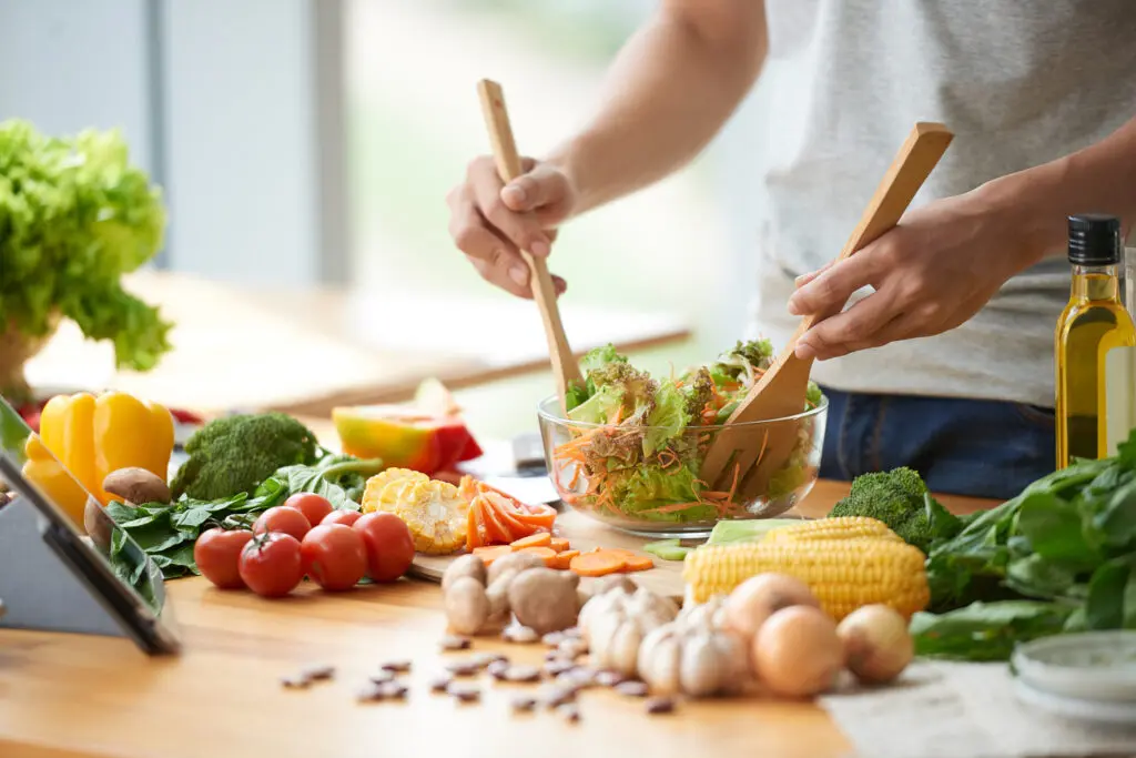 Photo of cooking ingredients on a worktop with a person stirring a glass salad bowl of salad. On the worktop there's garlic, onions, mushrooms, broccoli, spinach leafs, olive oil, sweetcorn, carrots, tomatoes, broccoli, yellow and red peppers, as well as herbs in plant pots. The image is brightly lit and colourful.