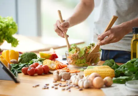 Photo of cooking ingredients on a worktop with a person stirring a glass salad bowl of salad. On the worktop there's garlic, onions, mushrooms, broccoli, spinach leafs, olive oil, sweetcorn, carrots, tomatoes, broccoli, yellow and red peppers, as well as herbs in plant pots. The image is brightly lit and colourful.
