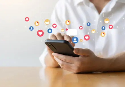 Close up of woman wearing a white blouse typing on her smartphone. There's cartoon images of 'Love', 'Like', 'Laugh', 'notification', and 'person' icons appearing to pop out of her phone.