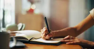 Photo of a young woman's hands and arms as she holds a pen while writing in a journal. The journal is lined, open, and has a dark, soft cover. She is holding a black pen in her right hand and is writing on the page. There's a coffee cup on the table and the background is blurred out. The image feels warm and soft. The girl also has silver jewellery on her right index finger and middle finger, and one on the middle finger of her left hand.