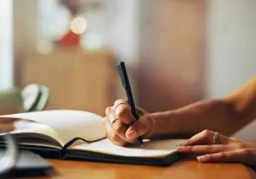 Photo of a young woman's hands and arms as she holds a pen while writing in a journal. The journal is lined, open, and has a dark, soft cover. She is holding a black pen in her right hand and is writing on the page. There's a coffee cup on the table and the background is blurred out. The image feels warm and soft. The girl also has silver jewellery on her right index finger and middle finger, and one on the middle finger of her left hand.