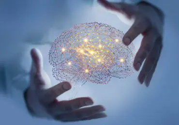 Image of a digital brain with connected lights inside it and two hands around it, but not touching it. The background is a blurred out light blue but we can slightly make out that the hands belong to a person.