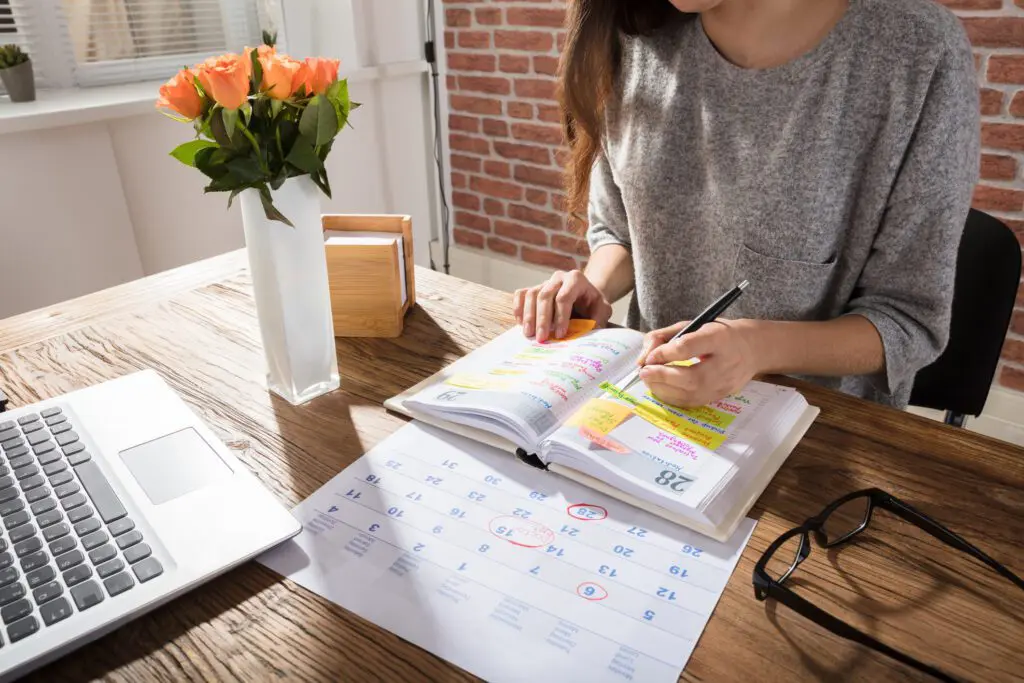 Woman writing colourful post it notes (she is left handed) and sticking them into a page in her diary. She's seated at a wooden table that also has her laptop, glasses, and a white vase containing orange roses. She is wearing a grey short-sleeved top and the photo cuts off at her chin - we only shows her chin and some brown hair hanging down.