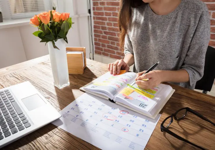 Woman writing colourful post it notes (she is left handed) and sticking them into a page in her diary. She's seated at a wooden table that also has her laptop, glasses, and a white vase containing orange roses. She is wearing a grey short-sleeved top and the photo cuts off at her chin - we only shows her chin and some brown hair hanging down.