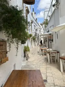 A typical street in Locorotondo, Italy. A narrow street with white buildings. Buildings are adorned with many green plants and flowers. There are a number of tables and chairs close to the wall on the right, belonging to a local restaurant.