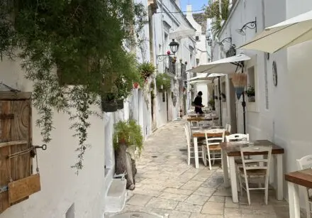 A typical street in Locorotondo, Italy. A narrow street with white buildings. Buildings are adorned with many green plants and flowers. There are a number of tables and chairs close to the wall on the right, belonging to a local restaurant.