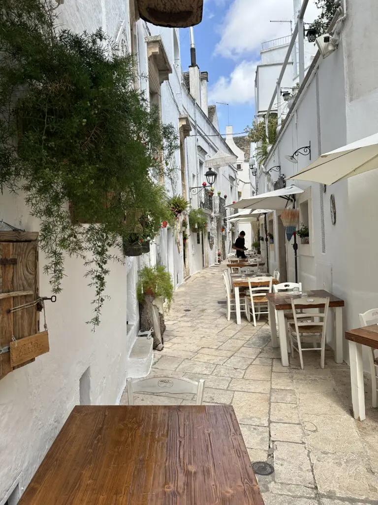 A typical street in Locorotondo, Italy. A narrow street with white buildings. Buildings are adorned with many green plants and flowers. There are a number of tables and chairs close to the wall on the right, belonging to a local restaurant.