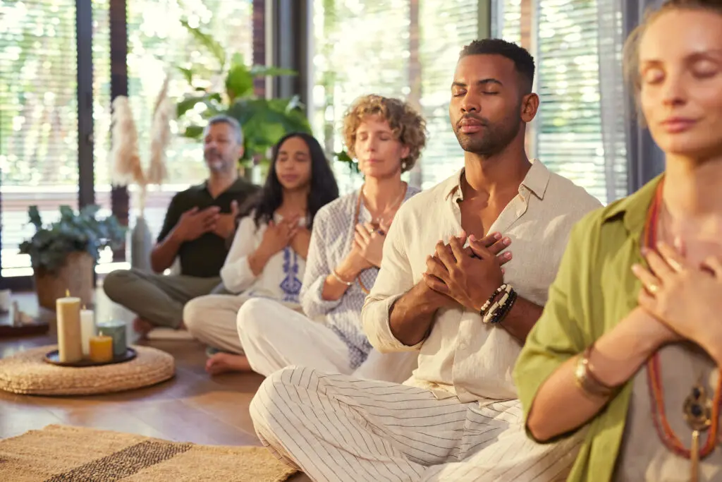 Photo of people meditating in a well lit meditation room. They all have their hands over their heart area. The room has some lit candles.