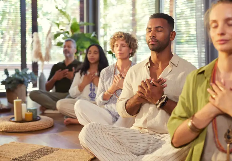 Photo of people meditating in a well lit meditation room. They all have their hands over their heart area. The room has some lit candles.