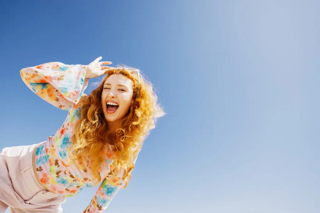 Happiness concept. Happy young woman with long orange curly hair, winking, laughing with radiant joy under a clear blue sky. She is wearing a pink flowery blouse and pink trousers and is leaning in from the side of the image.