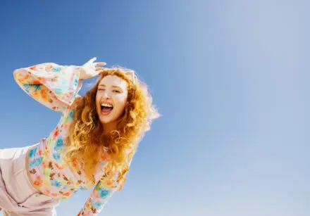 Happiness concept. Happy young woman with long orange curly hair, winking, laughing with radiant joy under a clear blue sky. She is wearing a pink flowery blouse and pink trousers and is leaning in from the side of the image.