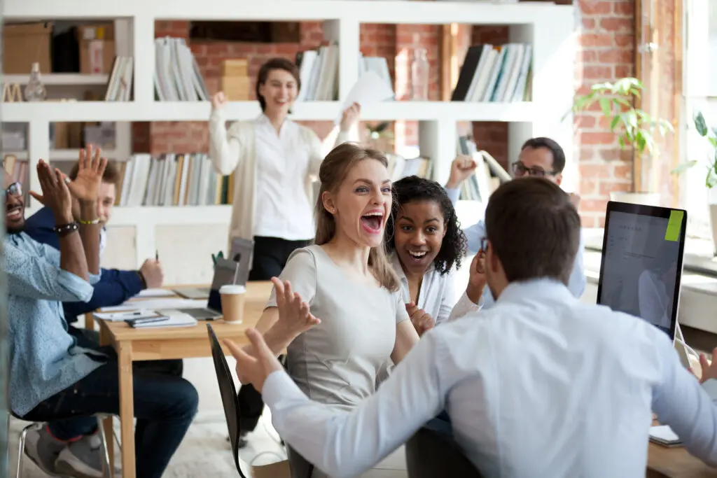 Capitalisation. Joy shared. Image of people in an open plan office celebrating one person's good news. They are smiling and cheering.
