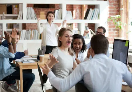 Capitalisation. Joy shared. Image of people in an open plan office celebrating one person's good news. They are smiling and cheering.