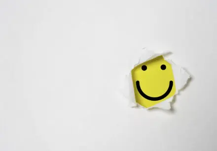 Happiness. Yellow smiley face peeking out through a hole in a piece of paper.