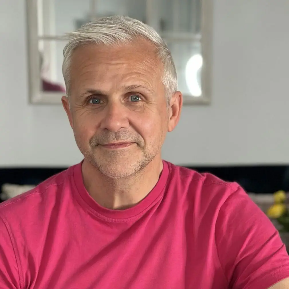 Dr David R Hamilton wearing pink t-shirt. Smiling. Pink flowers to his right, yellow flowers to his left.