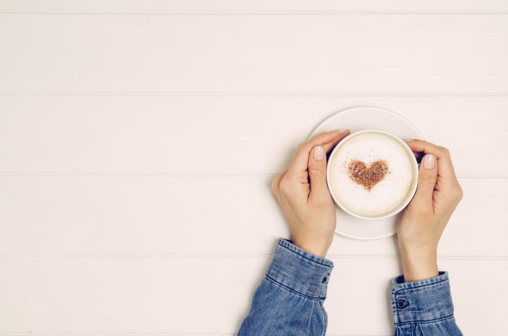 Female hand holding cup of coffee on white wooden table. The coffee has a heart made out of chocolate dust on it.