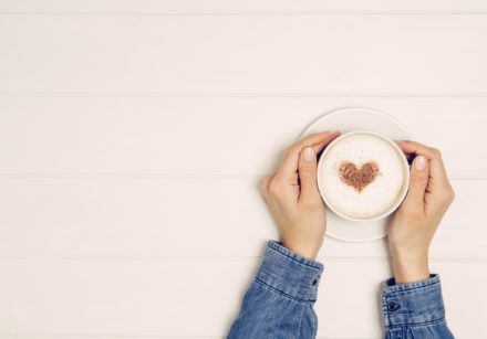 Female hand holding cup of coffee on white wooden table. The coffee has a heart made out of chocolate dust on it.