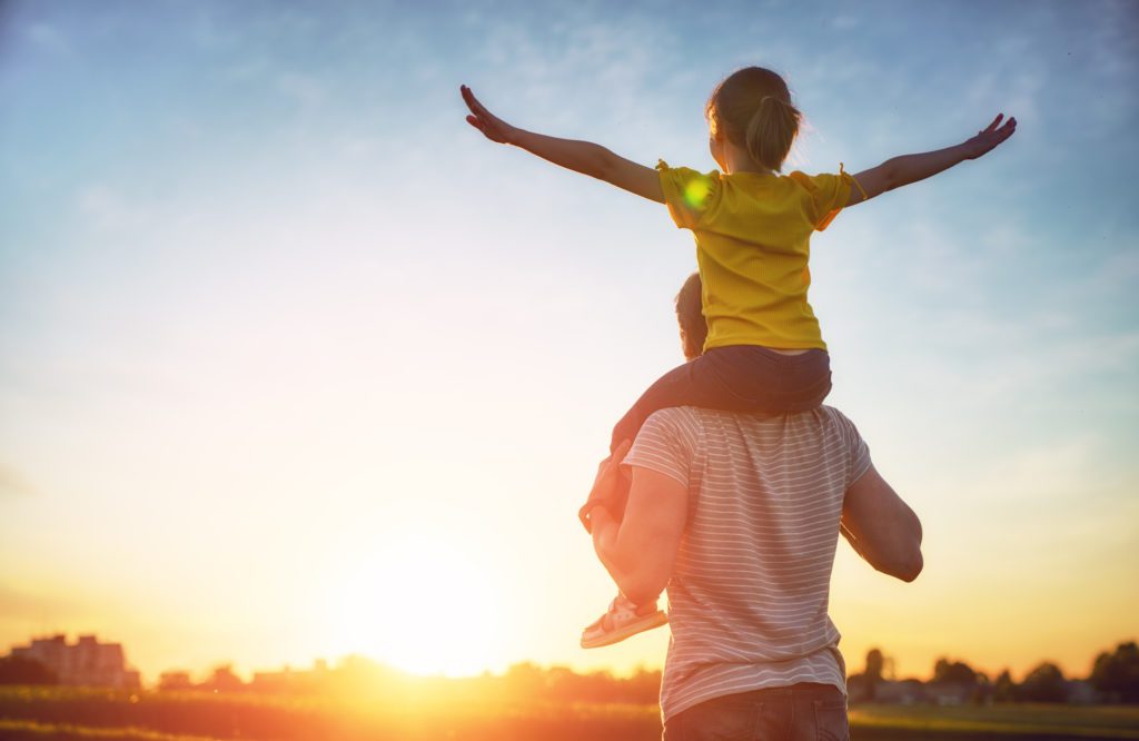 Father and his child playing together. Child on father's shoulders with arms outstretched, looking at a sunset.