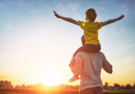 Father and his child playing together. Child on father's shoulders with arms outstretched, looking at a sunset.