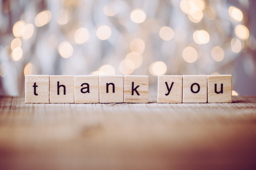 thank you spelled out in wooden blocks on a table, with soft white lights in the background.