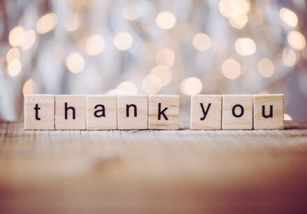 thank you spelled out in wooden blocks on a table, with soft white lights in the background.