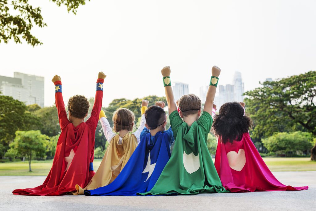 Five children wearing colourful capes, facing out onto a dark with buildings in the distance. They have their hands in the air expressing power, like they're superheroes.