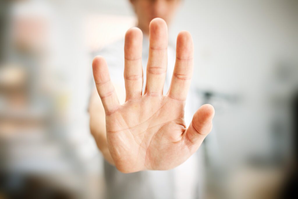 Man showing stop gesture with his hand out front. His face and body are blurred in the background and only the hand out in front is in focus.