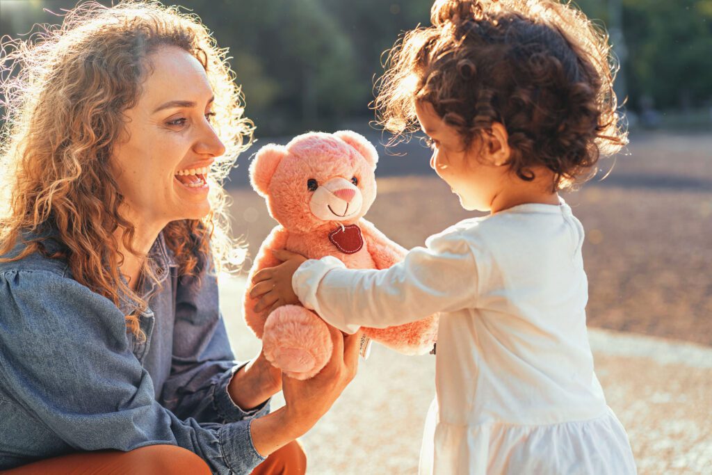 Curly haired woman giving a teddy bear to her little daughter outdoors in the park - family lifestyle concept.