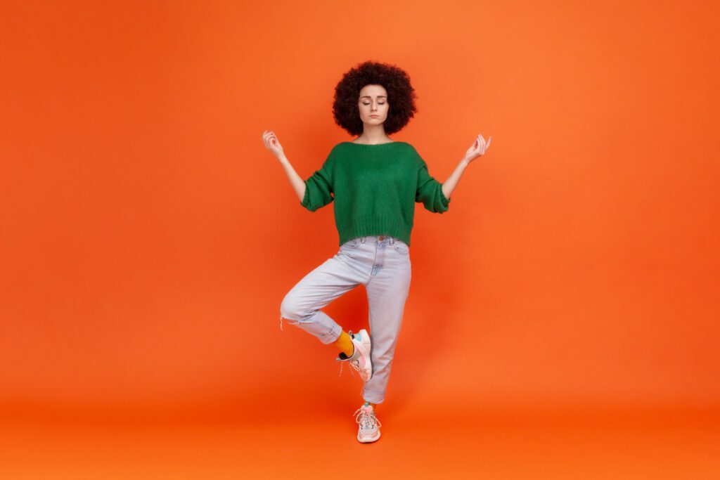 Portrait of woman with Afro hairstyle wearing green sweater with mudra gesture hands up, closed eyes, meditating standing in yoga position. Indoor studio shot isolated on orange background.